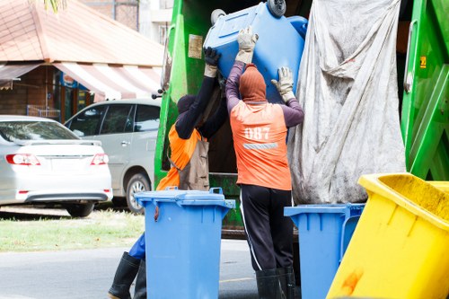 Two waste disposal workers wearing high-visibility orange vests and protective gloves are lifting a large blue plastic barrel into the back of a green rubbish collection vehicle parked on a residential street. One worker's head is covered with a hood, while the other is wearing a beanie. The vehicle has a grey tarpaulin partially covering the open rear compartment. In the foreground, three plastic bins – two blue and one yellow – are positioned on the pavement, with the yellow bin slightly tilted. Behind the scene, a row of parked cars, including a white vehicle, is visible along the curbside, with suburban houses and trees in the background. The environment suggests a typical UK residential neighbourhood, possibly within the postcode area associated with Belgravia, and the scene captures a routine rubbish collection activity handled by Belgravia House Clearance as part of their waste removal services.