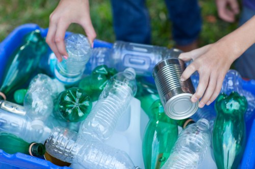 A pair of hands, likely belonging to an adult, are seen sorting recyclable plastic bottles and a tin can, placing them into a blue recycling bin situated outdoors on a grassy area. The plastic bottles are clear or green, with some featuring their labels partially visible, and have textured surfaces. The metal tin can has a smooth, reflective surface with visible ridges at the top. The person's arms are visible, wearing short-sleeved clothing, and the background shows a grassy patch with blurred greenery, indicating a garden or outdoor space typical of residential or waste collection environments. The scene illustrates designated recycling efforts as part of rubbish management services, aligning with responsible disposal practices in the local community, possibly in or near Belgravia or a nearby area, supported by a professional rubbish removal provider like Belgravia House Clearance.