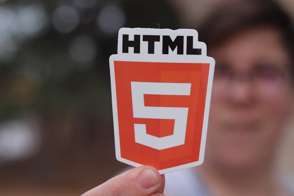 A person holding a cutout in front of their face displaying the HTML5 logo, which consists of a shield-shaped emblem with a bold, white number '5' on an orange background and the text 'HTML' in black