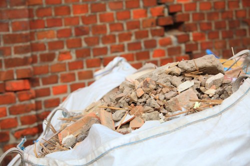 A close-up view of a large white construction waste sack filled with rubble, including broken bricks, concrete fragments, and small pieces of plaster, placed on a paved outdoor surface near a red brick wall. The rubble appears freshly collected with uneven textures and muted earthy tones, contrasting with the smooth, white fabric of the sack. In the background, parts of the brick wall and additional debris are visible, indicating a site in need of rubbish removal services. The scene is well-lit, suggesting daytime conditions typical of a professional clearance operation by Belgravia House Clearance, serving areas around Belgravia and central London.