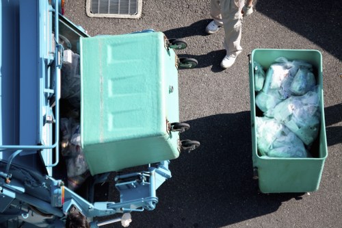 An overhead view of a light green, wheeled rubbish bin attached to a waste collection vehicle on a dark tarmac driveway. The bin is partially open, revealing a collection of assorted plastic bags, some containing waste and others appearing to be discarded packaging, with the bags showing various shades of white, blue, and transparent materials. To the right, there is another similar light green container filled with rubbish, also containing plastic bags and waste materials. A portion of a person wearing light-colored trousers and white footwear is visible in the background, standing next to the vehicle. The setting is outdoors, likely on a residential street or driveway, with natural lighting indicating daytime. The presence of the vehicle and waste materials is consistent with rubbish removal services in the local area, such as Belgravia or nearby districts, supporting the services offered by Belgravia House Clearance.